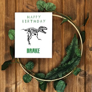Puede incluir: Una tarjeta blanca con texto verde que dice "Feliz cumplea&ntilde;os" y una ilustraci&oacute;n de esqueleto de dinosaurio en blanco y negro. La tarjeta tambi&eacute;n tiene el nombre "Drake" escrito en verde.