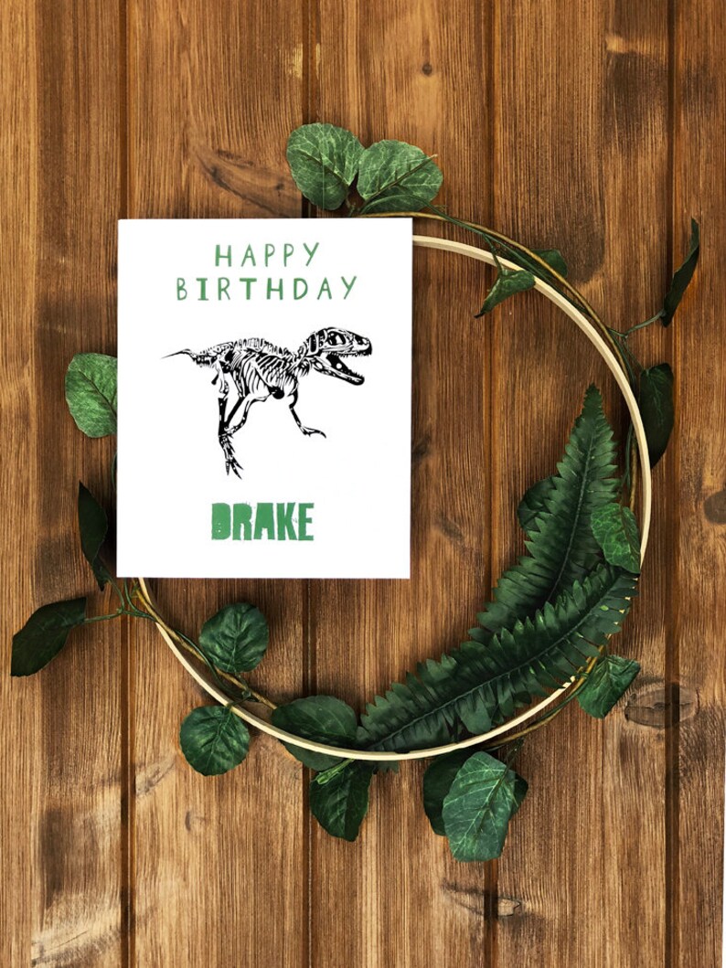 Puede incluir: Una tarjeta blanca con texto verde que dice "Feliz cumplea&ntilde;os" y una ilustraci&oacute;n de esqueleto de dinosaurio en blanco y negro. La tarjeta tambi&eacute;n tiene el nombre "Drake" escrito en verde.