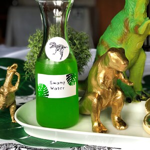 Puede incluir: Una botella de vidrio transparente con un l&iacute;quido verde en su interior. La botella tiene una etiqueta que dice "Swamp Water" y una pegatina con un esqueleto de dinosaurio. La botella est&aacute; sobre una bandeja blanca con una figura de dinosaurio dorada y una figura de dinosaurio verde.