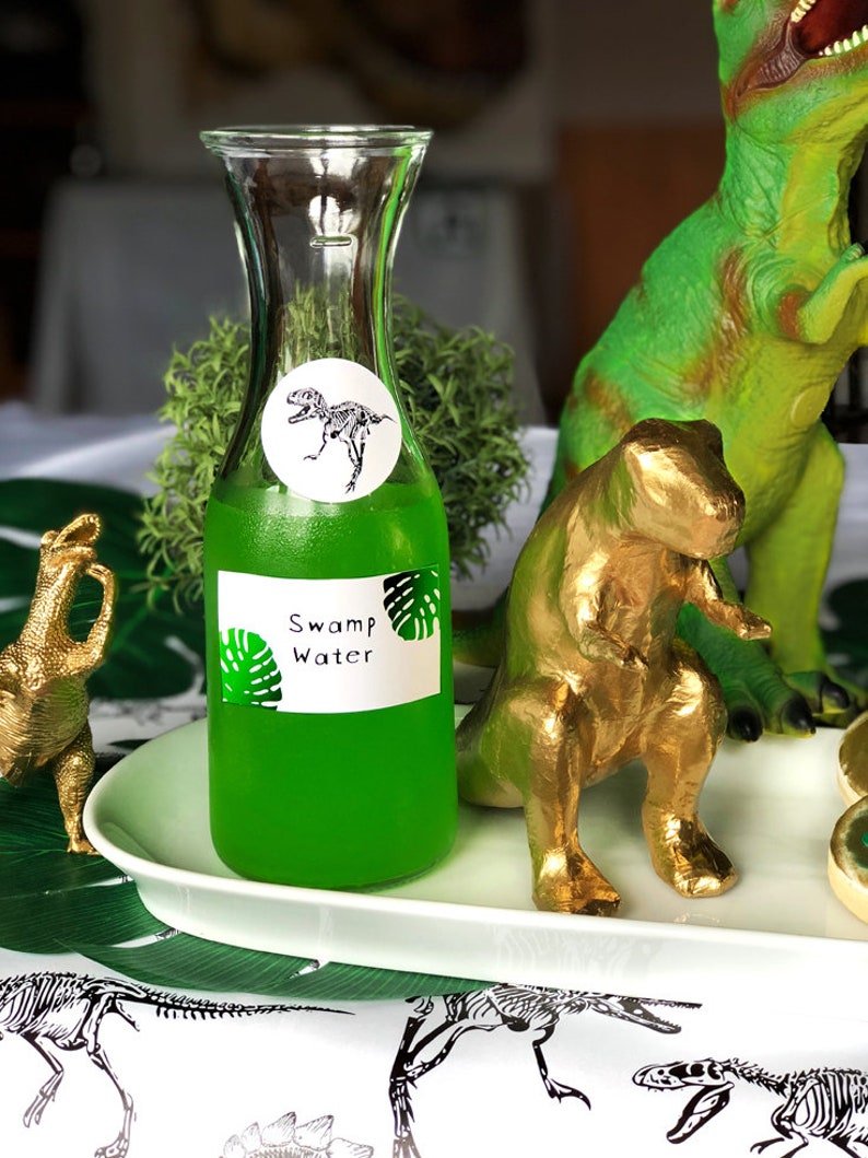 Puede incluir: Una botella de vidrio transparente con un l&iacute;quido verde en su interior. La botella tiene una etiqueta que dice "Swamp Water" y una pegatina con un esqueleto de dinosaurio. La botella est&aacute; sobre una bandeja blanca con una figura de dinosaurio dorada y una figura de dinosaurio verde.