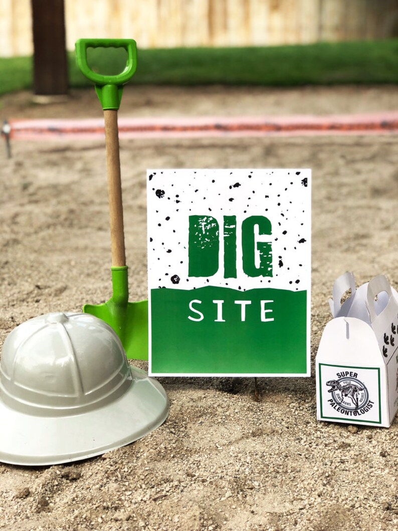 Puede incluir: Una pala de juguete de pl&aacute;stico verde con mango de madera, un cartel blanco y verde que dice "DIG SITE", un sombrero blanco de ala ancha y una caja blanca con un logotipo de dinosaurio verde que dice "Super Paleont&oacute;logo".