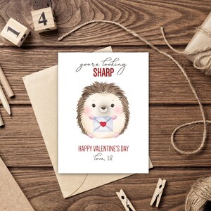 Puede incluir: Una tarjeta de San Valentín con un erizo de acuarela sosteniendo una carta en forma de corazón. La tarjeta dice "You're looking sharp" y "Happy Valentine's Day, Love, Liz" sobre un fondo de madera con lápices, cuerda y un regalo.