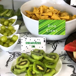 Puede incluir: Una mesa preparada para una fiesta con tem&aacute;tica de dinosaurios con uvas verdes, rodajas de kiwi y un bol de aperitivos de conos de ma&iacute;z amarillos. La mesa est&aacute; cubierta con un mantel blanco con un patr&oacute;n de esqueleto de dinosaurio negro. Los aperitivos est&aacute;n etiquetados con carteles que dicen "Garras de Dino" y "Cola de Brachiosaurus".