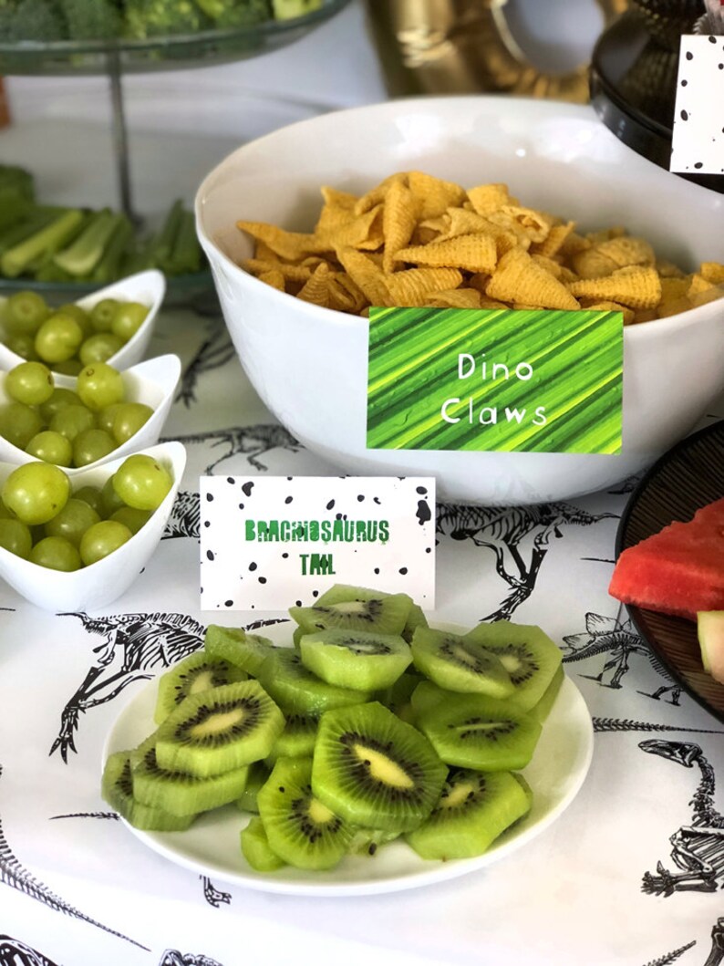 Puede incluir: Una mesa preparada para una fiesta con tem&aacute;tica de dinosaurios con uvas verdes, rodajas de kiwi y un bol de aperitivos de conos de ma&iacute;z amarillos. La mesa est&aacute; cubierta con un mantel blanco con un patr&oacute;n de esqueleto de dinosaurio negro. Los aperitivos est&aacute;n etiquetados con carteles que dicen "Garras de Dino" y "Cola de Brachiosaurus".