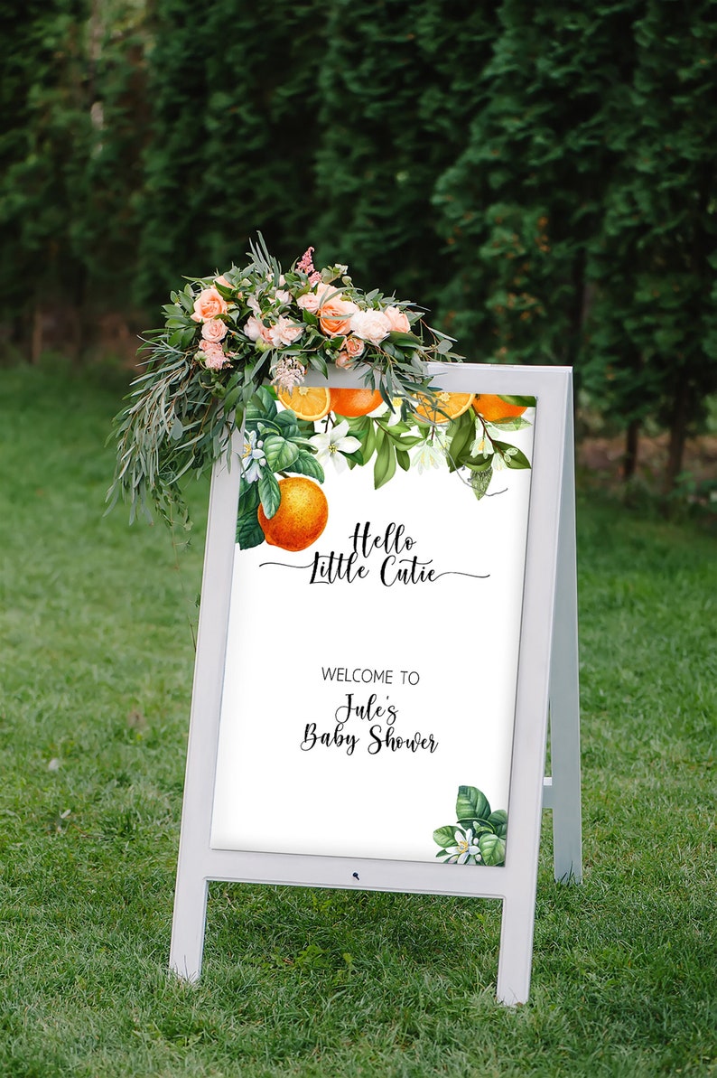 Puede incluir: Un letrero blanco en forma de A con el texto "Hello Little Cutie" y "Welcome to Jules Baby Shower". El letrero est&aacute; decorado con ilustraciones bot&aacute;nicas de naranjas y hojas verdes, coronado con un arreglo floral de flores rosas y melocot&oacute;n.