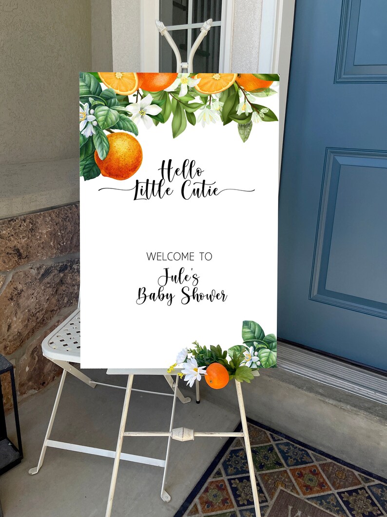 Puede incluir: Un letrero blanco sobre un caballete blanco con el texto "Hello Little Cutie" y "Welcome to Julie's Baby Shower". El letrero est&aacute; decorado con naranjas de acuarela, hojas verdes y flores blancas. El caballete est&aacute; sobre una alfombra estampada.