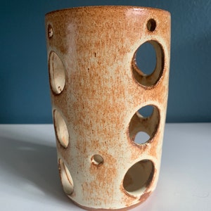 May include: A white ceramic planter with brown speckles and multiple circular cutouts. The planter is cylindrical in shape.