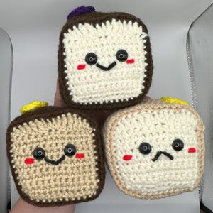May include: Three crocheted bread plush toys with different facial expressions. The bread is brown with a white crocheted centre. The bread has yellow and purple accents.