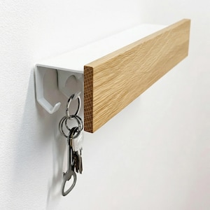 Könnte beinhalten: Ein Wand-Schlüsselhalter mit einem weißen Metallregal und einem Akzent aus Naturholz. Das Regal hat einen Haken für Schlüssel und eine Holzfront. Ein Schlüsselbund hängt am Haken. Das Design ist minimalistisch und modern.