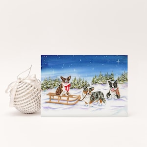 May include: A Christmas card featuring a watercolor painting of three corgi dogs in a snowy winter scene. One dog is on a wooden sled, and the others are standing in the snow. A silver ornament is to the left.