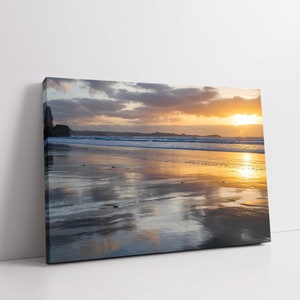 May include: A scenic photograph of a beach at sunset. The sky is a mix of orange, pink, and purple, with clouds reflecting in the water. The sand is wet and glistening, with the waves gently rolling in.