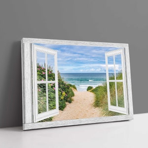 May include: A white window frame with two open windows, revealing a view of a sandy path leading to a beach with blue ocean waves. The path is lined with green grass and bushes.
