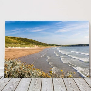 May include: A coastal print showing a sandy beach, green hills, and a clear blue sky with scattered clouds. Waves gently break on the shore. The print is displayed on a white wall with a wooden floor.