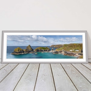 May include: A panoramic photograph of a rocky coastline with a small beach and a few white houses. The water is a vibrant turquoise blue and the sky is a clear blue with white clouds.