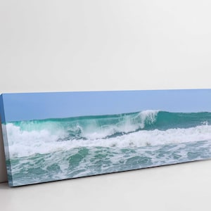 May include: A panoramic photograph of a large ocean wave breaking on the shore. The wave is a vibrant turquoise colour with white foam cresting at the top. The water is a deep blue and the sky is a light blue.