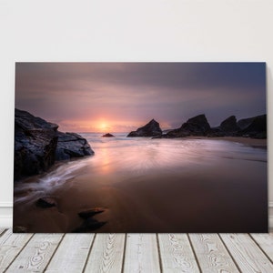 May include: A scenic photograph of a rocky beach at sunset. The sky is a vibrant purple and pink, with the sun setting behind the rocks. The water is calm and reflects the colours of the sky.