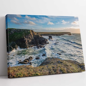 May include: A scenic landscape photograph of a rocky coastline with crashing waves and a blue sky with white clouds. The sun is setting in the distance, casting a warm glow on the scene.