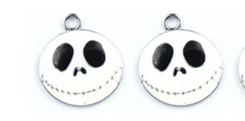 Op de afbeelding: Ronde, witte emaille bedels met een zwart-wit gezichtsontwerp en een gestikte glimlach. Elke bedel heeft een zilverkleurige lus aan de bovenkant om aan sieraden of andere knutselwerken te bevestigen. Het ontwerp doet denken aan een populair animatiefiguur.