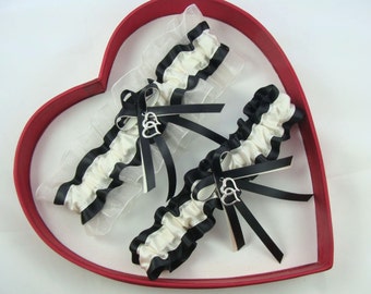 May include: Two white and black garters with silver double heart charms. The garters are displayed in a red heart-shaped box.