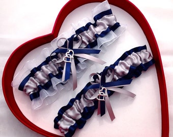 May include: Two white and silver garters with navy blue satin ribbon and a silver double heart charm. The garters are displayed in a red heart-shaped box.