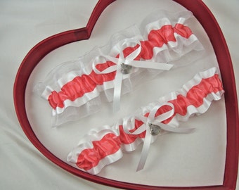 May include: Two white and coral garters with a silver charm in the shape of a seashell. The garters are displayed in a red heart-shaped box.
