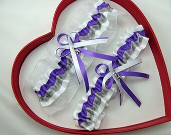 May include: Two white and purple garters with silver charms. The garters are made with white satin ribbon, purple satin ribbon, and white tulle. The charms are shaped like hearts with the word "love" on them.