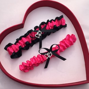 Könnte beinhalten: Zwei rosa und schwarze Strumpfhalter mit silbernen Doppelherzen. Die Strumpfhalter werden auf einer roten herzförmigen Schachtel präsentiert.