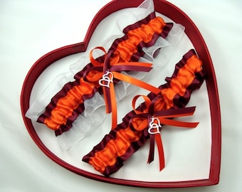 May include: Two wedding garters with orange and burgundy ruffles, accented with silver heart charms and ribbons. The garters are displayed in a heart-shaped box with a red rim and white interior.