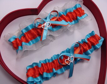 May include: Two turquoise and orange garters with silver heart charms. The garters are on a red heart-shaped box.