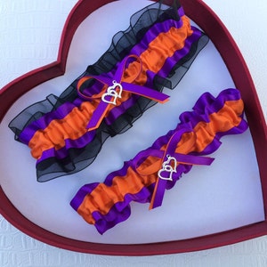 May include: Two bridal garters with black, orange, and purple satin ribbon and a silver double heart charm.