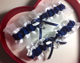 May include: Two white and navy blue garters with silver anchor charms. The garters are displayed in a red heart-shaped box.