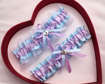 May include: Two blue and purple satin garters with a silver lock and key charm. The garters are displayed in a red heart-shaped box.