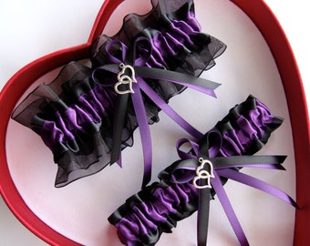 May include: Two purple and black garters with silver double heart charms. The garters are made with satin ribbon and black lace.