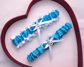 May include: Two wedding garters. One garter is turquoise with white ruffled trim and a white bow. The other garter is turquoise with a white bow. Both have silver-colored charms.