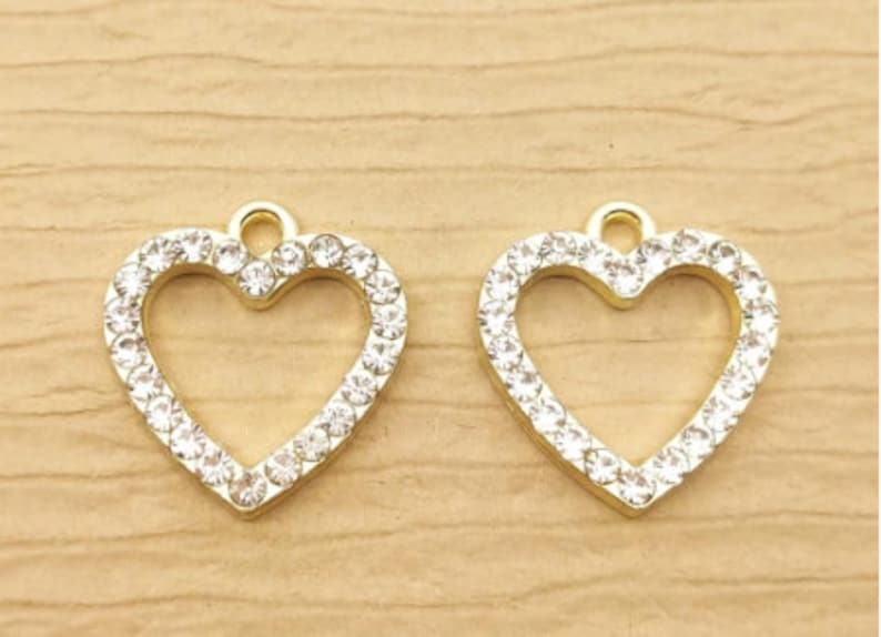 Op de afbeelding: Twee goudkleurige hartvormige bedels met heldere kristallen accenten. Elke bedel heeft een lus aan de bovenkant om op te hangen. De kristallen zijn rond de omtrek van het hart gezet. De achtergrond is een lichte houtnerf.
