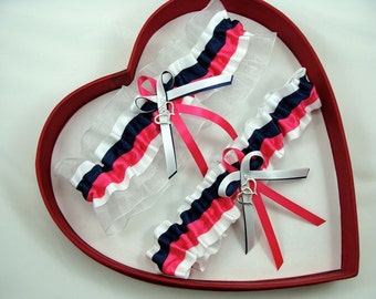 May include: Two white, pink, and navy blue garters with silver heart charms. The garters are displayed in a red heart-shaped box.