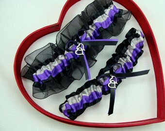 May include: Two wedding garters with black, silver, and purple satin and organza ruffles. Each garter has a silver heart charm and black ribbon. The garters are displayed inside a red heart-shaped frame.