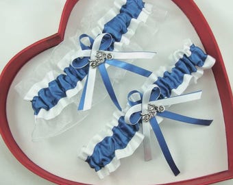 May include: Two white and blue garters with a silver charm in the shape of a heart with wings. The garters are in a red heart-shaped box.
