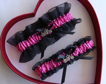 May include: Two black and hot pink garters with a silver motorcycle charm. The garters are displayed in a heart-shaped box.