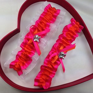 Könnte beinhalten: Zwei weiße und rosafarbene Strumpfhalter mit orangefarbenem Satinband und einem silbernen Schmetterlingsanhänger. Die Strumpfhalter werden in einer roten herzförmigen Schachtel präsentiert.