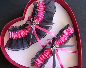 May include: Two pink and black garters with a silver motorcycle charm. The garters are displayed in a red heart-shaped box.