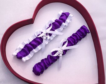 May include: Two purple and white satin garters with silver heart charms. The garters are displayed in a heart-shaped box.