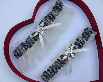 May include: Two white bridal garters with a camouflage pattern and a silver deer charm. The garters are displayed on a red heart-shaped box.
