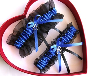 May include: Two blue and black garters with silver double heart charms. The garters are in a red heart-shaped box.