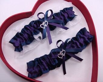 May include: Two purple and navy blue garters with white lace and silver double heart charms. The garters are displayed in a red heart-shaped box.