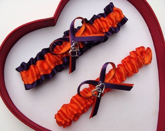 May include: Two orange and purple satin garters with silver heart charms. The garters are on a red heart-shaped background.