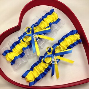 May include: Two blue and yellow striped garters with white lace and silver heart charms. The garters are displayed in a red heart-shaped box.
