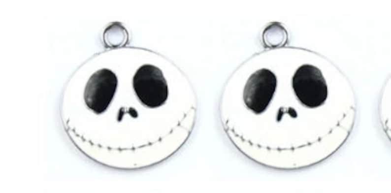 Puede incluir: Dos abalorios de metal plateado con fondo de esmalte blanco y detalles de esmalte negro. Cada abalorio presenta una cara inspirada en Jack Skellington con una sonrisa cosida.