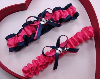 May include: Two pink and navy blue garters with a silver heart and key charm. The garters are on a red heart-shaped background.