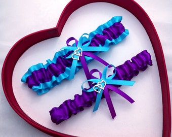 May include: Two bridal garters with purple and turquoise satin ribbon and a silver charm with three hearts. The garters are displayed in a red heart-shaped box.
